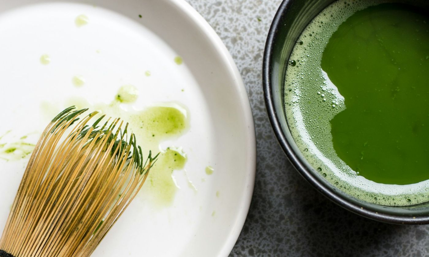 A bamboo tea whisk and a bowl of whisked green matcha on a grey stone surface